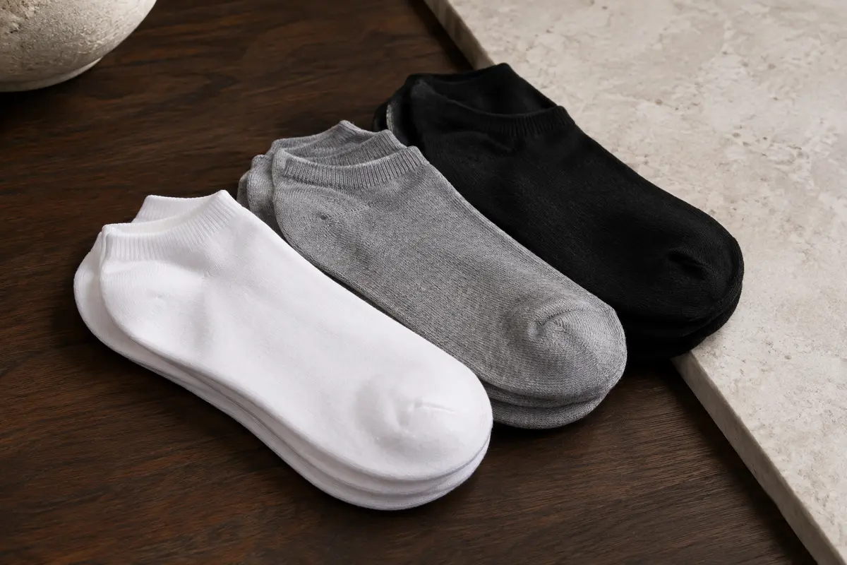 Socquettes basses unisexes présentées en blanc, noir et gris sur du bois foncé et de la pierre claire.