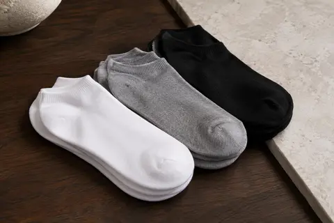 Socquettes basses unisexes présentées en blanc, noir et gris sur du bois foncé et de la pierre claire.